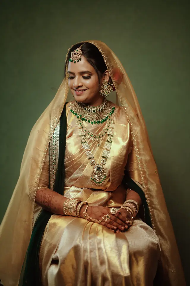 Bride in a gold tissue dupatta with an emerald and gold necklace, makeup by Popyz