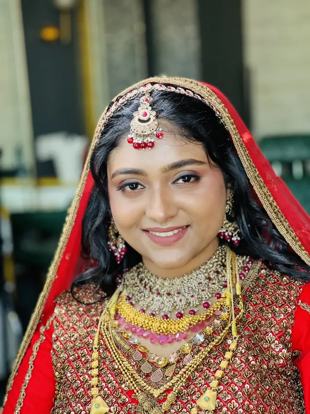 Close-up of a smiling bride wearing a red embroidered dupatta over her head, layered gold necklaces and a gold maang tikka