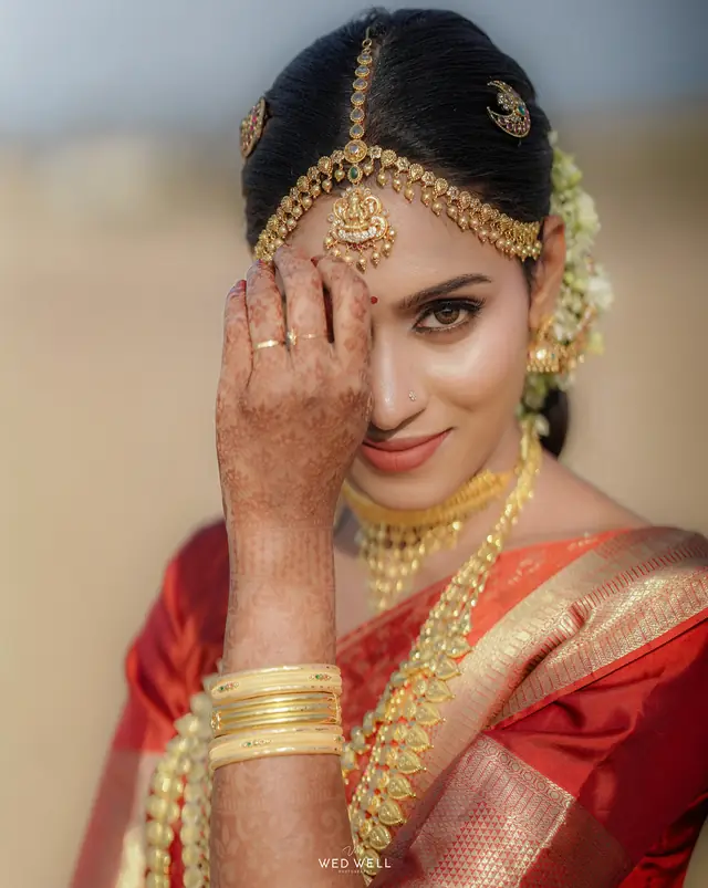 Bride outdoors in soft light wearing a red-and-gold silk saree, layered temple-style gold jewellery and jasmine flowers in her hair
