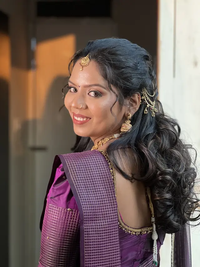 A woman with a warm deep skin tone and soft curls, wearing a purple silk saree and glowing makeup in warm evening light