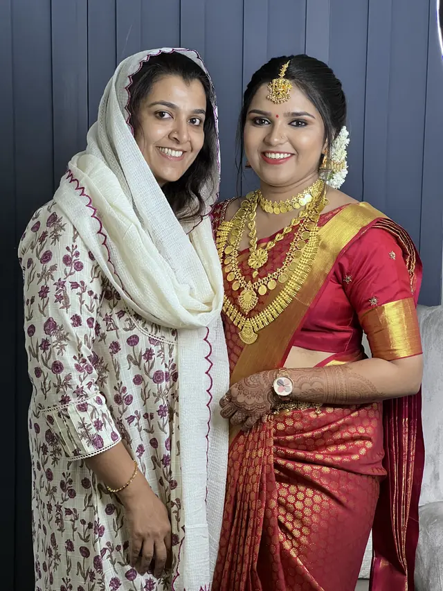 Bride in a red and gold silk saree standing beside a guest in a white scarf