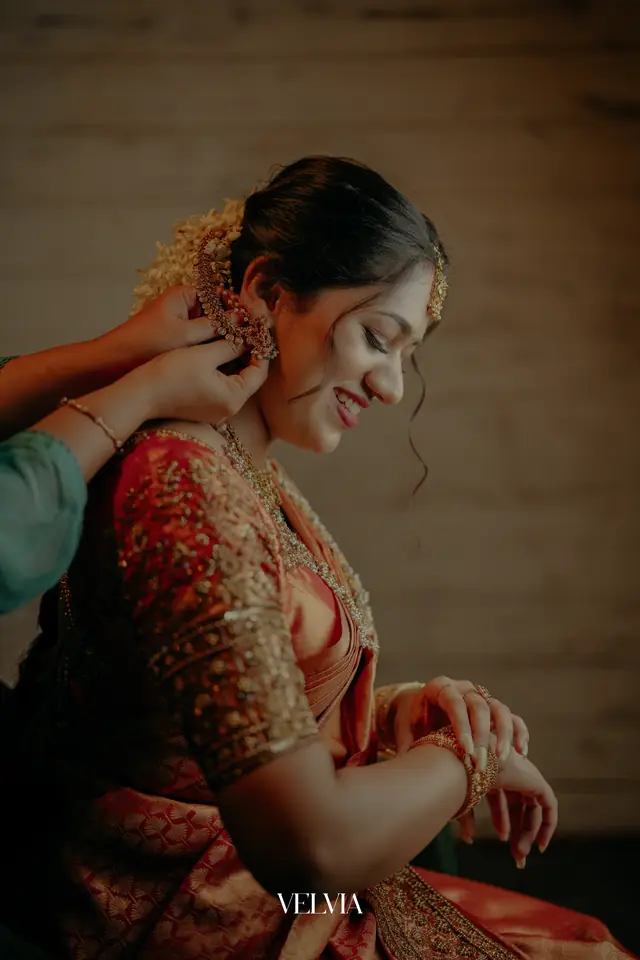 Bride in a red silk saree having her jewellery fastened before the ceremony