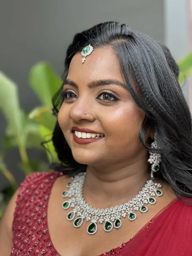 Close-up of a smiling woman with dewy skin wearing a red outfit and an emerald-and-diamond necklace, with green plants behind her