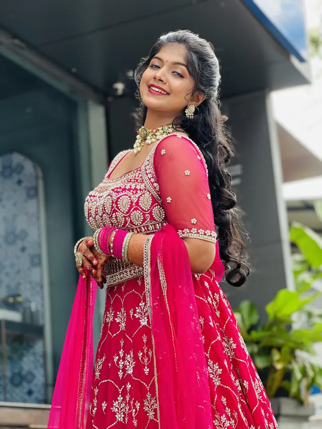 Young woman in a pink embroidered lehenga standing outdoors beside plants in front of a building, smiling, with fresh glowing makeup